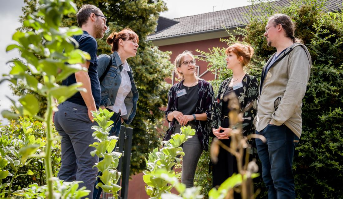 Fünf Personen stehen zwischen Hecken in lockerem Gespräch zusammen