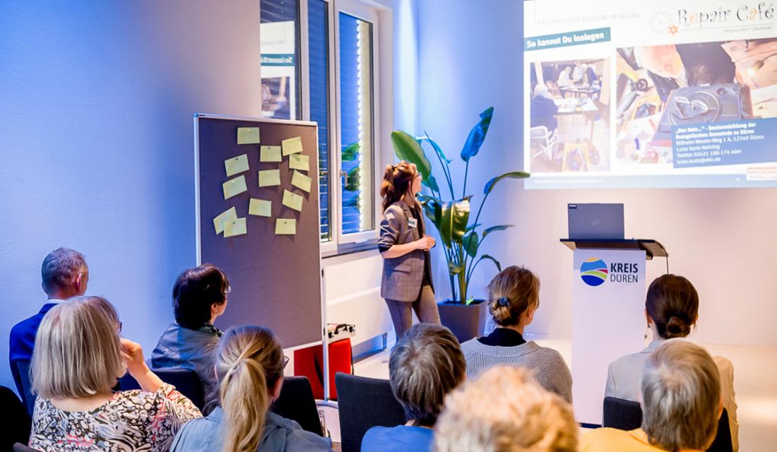 Aus dem Publikum fotografiert: Eine Frau hält einen Vortrag und blickt auf eine Leinwand hinter sich